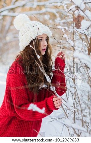 Young girl, brunette, with long hair posing on a sunny winter day. On it: a red sweater, a white hat. Around the winter forest and snow-covered branches.