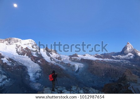 A tourist standing on the edge of a rocky ridge, taking photos of the iconic Matterhorn among snow capped alpine mountains illuminated by bright moonlight in Gornergrat, Zermatt, Valais, Switzerland