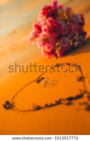 Cute wedding rings, flower and hear on the sand beach. Royalty high-quality free stock image of background romantic couple wedding rings in heart shape on the sand and bunch rose flowers. Card design