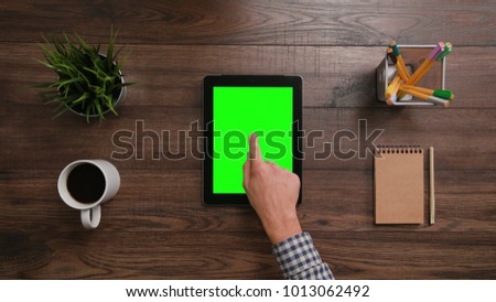 A man's finger touching an Digital Tablet with a green screen. The Digital Tablet is on the brown table. View from the top. Close-up.