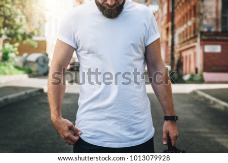 Summer day. Front view. Young bearded millennial man dressed in white t-shirt is stands on city street. Mock up. Space for logo, text, image. Instagram filter, film effect, bokeh effect.