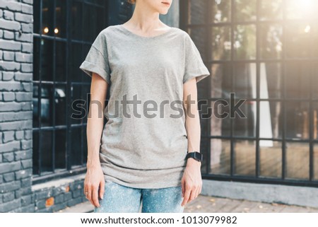 Summer day. Front view. Young millennial woman dressed in gray t-shirt is stands against gray wood wall. Mock up. Space for logo, text, image.