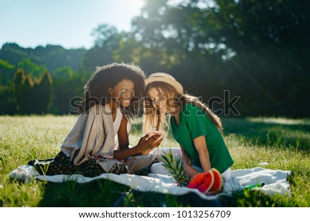 Charming african girl with pretty smile is showing something funny on a phone to her caucasian blonde girlfriend during the picnic on the sunny meadow.
