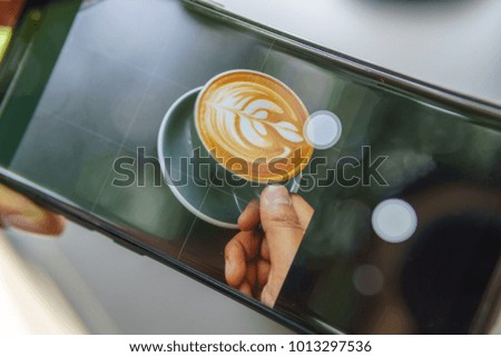 close up of smartphone taking photo of hand holding cup of latte art coffee.