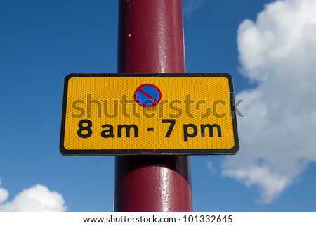 An oblong yellow sign with black border, the symbol for no parking and the time for no parking attached to a red post against a blue cloudy sky.