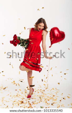 Full length portrait of a delighted young woman dressed in red dress holding bouquet of roses and air balloons while standing under falling confetti and celebrating isolated over white background