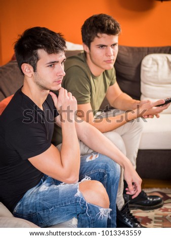 Two young friends lounging on sofa and watching TV or a movie with interest while chatting.