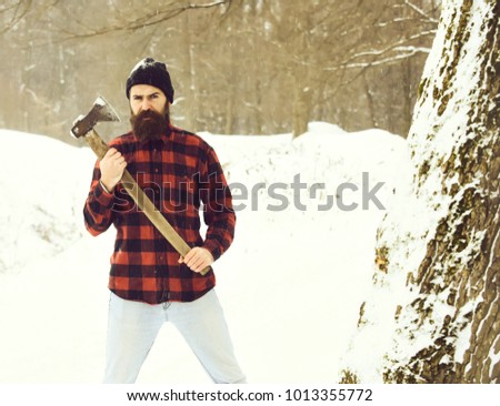 Handsome man or lumberjack, bearded hipster, with beard and moustache in red checkered shirt stands with axe in snowy forest on winter day outdoors on natural background