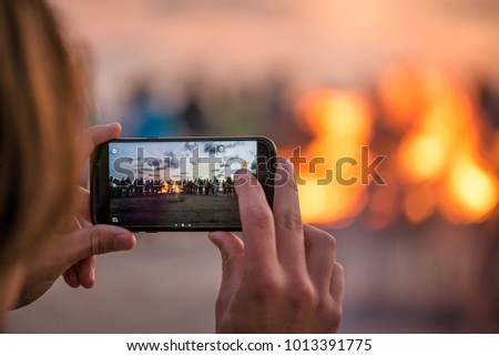 Woman is taking picture of sunset with smart phone. Romantic bonfire night at seaside. People gathering together to celebrate Night of ancient lights. Large burning campfire with soft glowing flame
