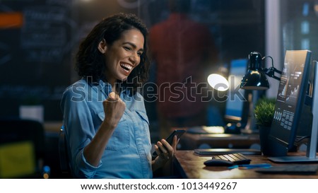 Beautiful Female Financier Uses Mobile Phone, Has Great News and Shows "Yes" Gesture, Her Personal Computer Showing Graphs and Statistics. She Smiles Charmingly. Royalty-Free Stock Photo #1013449267