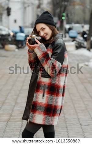 Girl walking and taking photos with her retro camera. Winter hipster look of plaid jacket and black beanie.