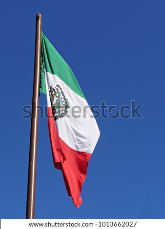Close up of the flag of Mexico unfurled and hanging down in the air against a backdrop of blue skies