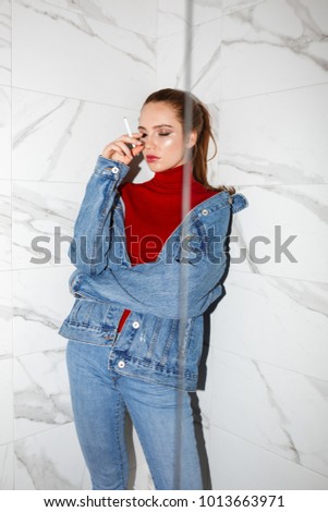Portrait of young beautiful woman in denim jacket and jeans standing with cigarette in hand and dreamily closing her eyes isolated