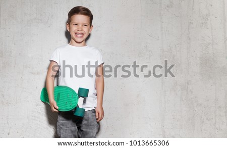 Cute smiling guy with skateboard in hand against gray wall. Portrait of skater boy in white T-shirt, concrete wall on background with copy space.