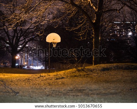 Nighttime photograph after dark of a white metal basketball backboard and orange hoop rim and white net near Foster Beach in Chicago in winter with black night background and trees framing the picture