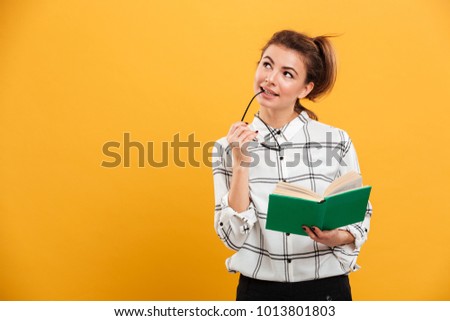 Photo of young brooding woman looking aside while holding book and eyeglasses in hands isolated over yellow background