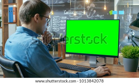Young Man Works at His Desk on the Personal Computer with Mock-up Green Screen. In the Background His Colleague Works in the Creative Office.