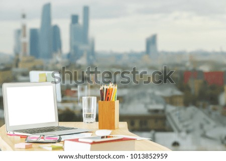 Close up of modern desktop with empty white laptop, supplies and other items on blurry city view background. Mock up 