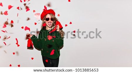 Lovely caucasian girl holding her shoes under heart confetti. Glad curly lady in green sweater enjoying valentine's day. Place for text