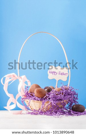 Image of chicken, chocolate eggs, purple decorative paper in basket on empty blue background with wish for happy Easter