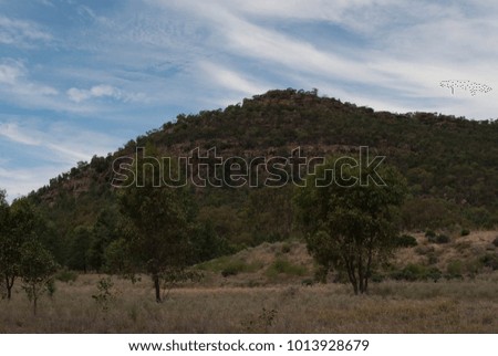 a large hill in the countryside with many trees on it and in foreground with clouds in the sky
