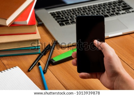 Man holding a phone, selective focus. Wooden desktop with a laptop, books and pens in the background. Mock-up for your picture.