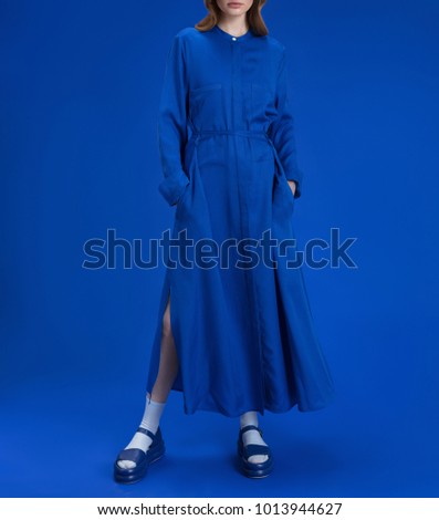 A young girl with blond long hair in a beautiful long blue dress shirt, in blue shoes, white socks on a blue background, studio shot