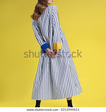 Young girl with long blonde hair in fashionable clothes, black and white with colored inserts long striped shirt dress posing on bright yellow background, studio shot