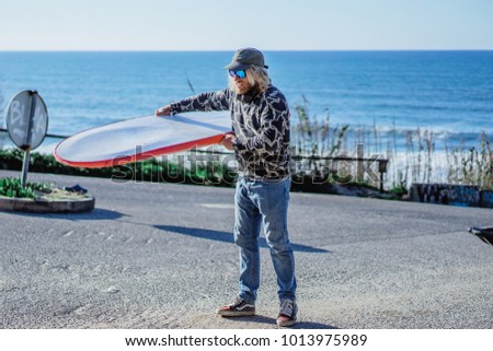 surfer man with long white hair in a cap and sunglasses with a surfboard. returns after surfing, folds the surfboard into the car. Nazare, Portugal.
