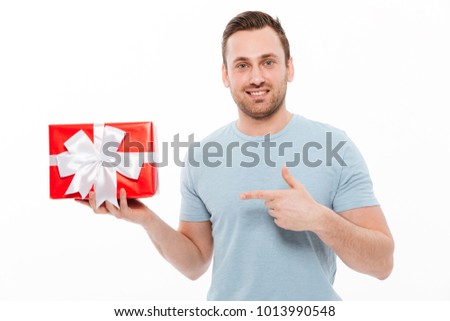 Image of good-looking guy having bristle smiling and pointing index finger on red gift box isolated over white background