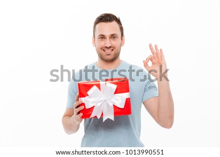 Image of pleased brunette guy smiling and showing OK sign while holding red present box with bow isolated over white background
