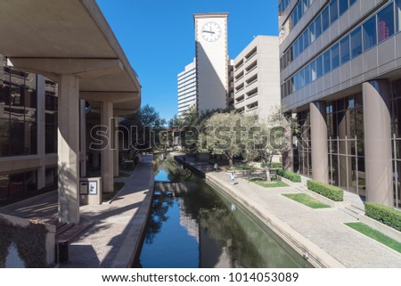 Clock tower and elevated apt light rail guide way alongside Mandalay Canal Walk in Las Colinas, Irving, Texas. Winding canal walkway along tree-lined path, people taking photo from cell phone.