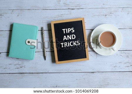 Top view of notebook,pen,a cup of coffee, and blackboard written with 'TIPS AND TRICKS' on white wooden background.