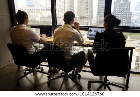 Behind of business man and woman sitting at the table and working with laptop in the office with window view of high building. The glass window view is at the front of the desk. The city window view.