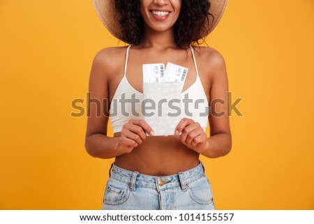 Close up portrait of a smiling african woman dressed in summer clothes holding passport with flying tickets isolated over yellow background