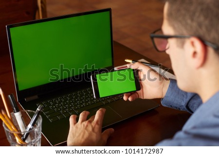 Back view of businessman using smartphone and laptop typing in office. Green screen mockup. Freelancer workspace. People and technology. Copyspace and product placement.