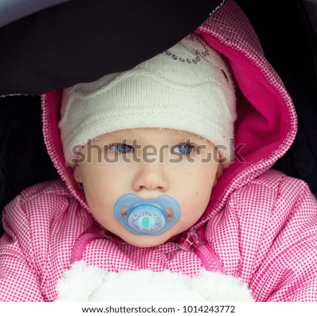 Little girl with teat in winter cap in pram, portrait, girl face.