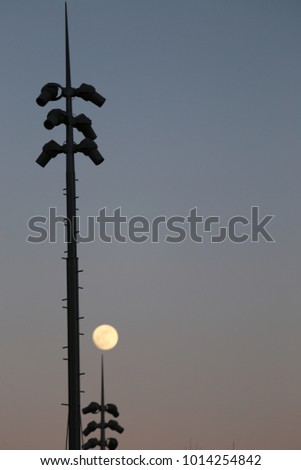 View of thin long vertical iron bars supporting spots. In background, the full moon in the purple sky. Strange composition of isolated elements. Abstract urban picture taken in winter in France.