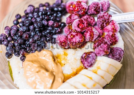 Macro closeup of oatmeal oat grain porridge bowl with sliced bananas, peanut butter, frozen berries, raspberries, blueberries, maple syrup