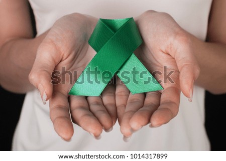 Close up of a slim woman in white t-shirt holding and showing green awareness ribbon in her hands on black background