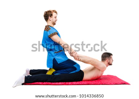 a young handsome man doctor masseur in a blue suit makes a traditional pose of takia massage to a guy lying on a living on a carpet rug. White isolated background. Theme Thai massage.