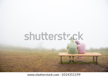 A couple who wear the rain cover suit sitting on the bamboo bench on the ground in the foggy atmosphere.