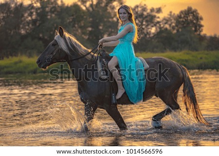 A girl riding running horse through shallow water of river at sunset background