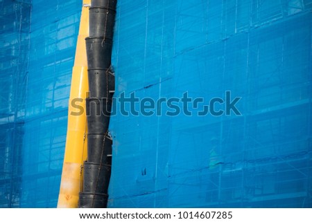Close up on yellow and black plastic garbage chutes on the exterior of an office building, with blue tarps covering scaffolding, at an industrial construction site