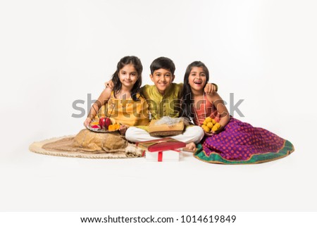 3 indian kids or siblings in traditional wear sitting on sofa or white background, holding gifts and sweets or laddu Royalty-Free Stock Photo #1014619849