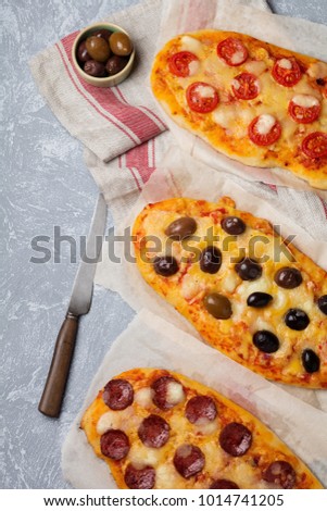 Three different oval pizza with olives, tomato and salami on a gray concrete background. Selective focus. Top view. Copy space.