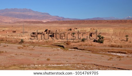 Traditional Moroccan fortified house on desert, dry river bed in foreground, few palms, in background blurry mountains with few tops covered with snow, sunny day, blue sky