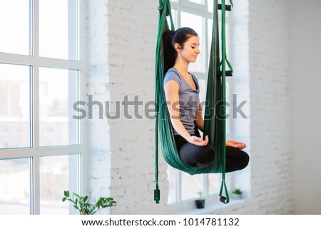 Woman sitting in lotus pose in hammock practicing aerial yoga in white loft interior studio. Fitness, sport, training and lifestyle concept.