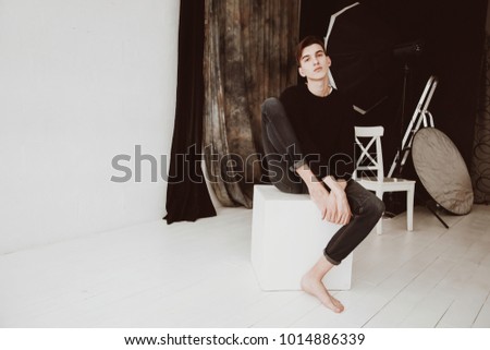 A handsome male model, in a photo studio. On the background of studio equipment. A guy barefoot, in jeans and a black sweater. Sits on a white cube.