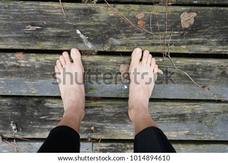 A pair of woman’s legs wearing black tight pants and standing barefoot on old textured wooden planks full of dry brown leaves and broken branches.